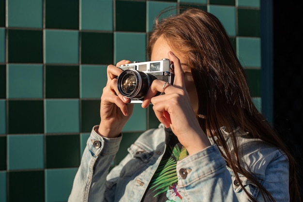 Cool meisjesmodel met retro filmcamera met een spijkerjasje, donker haar buiten over de stadsmuur op een kooiachtergrond. Plezier hebben in de stad met camera, reisfoto van fotograaf.