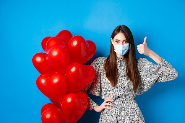 Coronavirus en pandemisch concept. Mooie vrouw met medisch masker en jurk in de buurt van Valentijnsdag ballonnen en duim opdagen, staande op blauwe achtergrond.