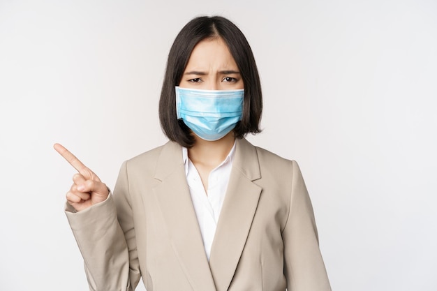 Coronavirus en werkconcept Gefrustreerde Aziatische zakenvrouw met medisch gezichtsmasker die met de vinger naar links wijst en klaagt over een witte achtergrond