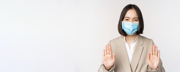 Coronavirus en werkconcept portret van een aziatische vrouwelijke kantoordame op de werkplek met een medisch gezichtsmasker en een stopverbodgebaar op een witte achtergrond