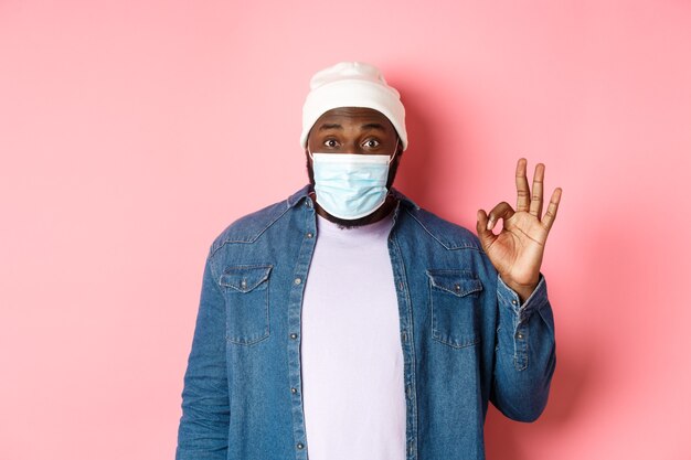 Coronavirus, levensstijl en sociaal afstandsconcept. Onder de indruk van een Afro-Amerikaanse man met een gezichtsmasker, met een goed teken, leuk en akkoord, staande over een roze achtergrond