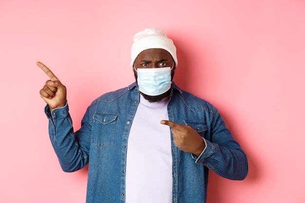 Coronavirus, levensstijl en wereldwijd pandemisch concept. Boze en teleurgestelde afro-amerikaanse man met gezichtsmasker naar links wijzend, starend naar camera ontevreden, roze achtergrond.