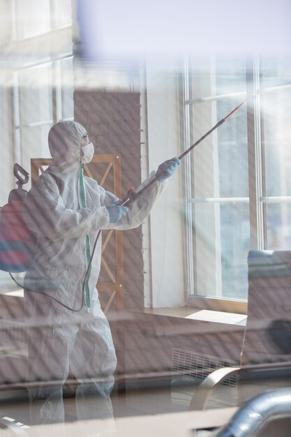 Coronavirus Pandemic. Een ontsmettingsmiddel in een beschermend pak en masker spuit ontsmettingsmiddelen in de kamer.