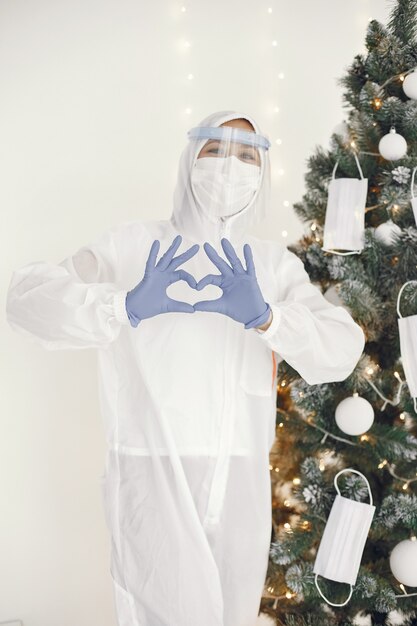 Coronavirus pandemie covid-2019. Beschermend pak, bril, handschoenen, masker. Kerstboom is versierd met medisch masker.
