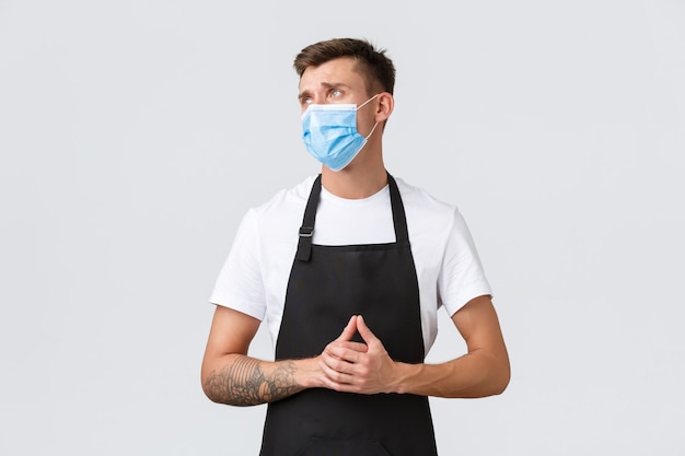 Coronavirus, sociale afstand in cafés en restaurants, zaken tijdens pandemisch concept. Teleurgestelde en verdrietige barista, verkoper in schort en medisch masker kijkt ontevreden en overstuur naar links