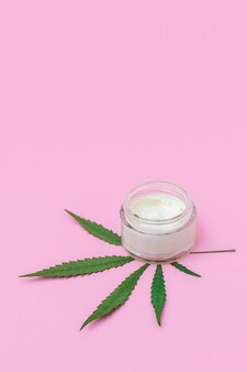 Cosmetische glazen pot met met cannabis doordrenkte gezichtscrème en groen marihuanablad op pastelroze achtergrond...