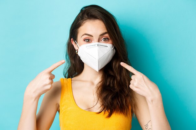 Covid-19, coronavirus en sociale afstand. Jonge vrouw in gasmasker wijzend op haar gezicht, vragend om gezichtsmaskers te gebruiken tijdens pandemie, staande tegen een blauwe achtergrond.