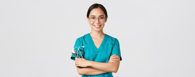 Covid-19, coronavirusziekte, concept van gezondheidswerkers. Close-up van zelfverzekerde professionele vrouwelijke arts, verpleegster in glazen en scrubs die op een witte achtergrond staan, kruisarmen.