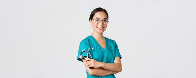 Covid-19, coronavirusziekte, concept van gezondheidswerkers. Professionele knappe Aziatische arts, medisch werker in glazen en scrubs, kruisarmen en glimlachen, witte achtergrond