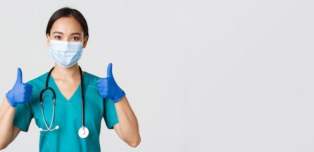 Covid-19, coronavirusziekte, concept van gezondheidswerkers. Zelfverzekerde glimlachende aziatische arts, verpleegster met medisch masker en rubberen handschoenen, duim omhoog in goedkeuring, service verlenen, witte achtergrond.