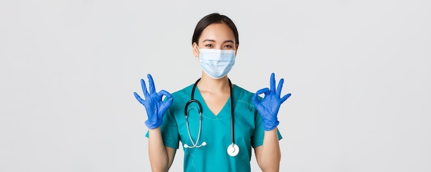 Covid-19, coronavirusziekte, concept van gezondheidswerkers. Zelfverzekerde glimlachende aziatische vrouwelijke arts, verpleegster met medisch masker en handschoenen, toont goed gebaar in goedkeuring, witte achtergrond.