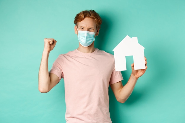 Covid-19 en onroerend goed concept. Gelukkig roodharige man in medisch masker, papier huis uitgesneden en vuist pomp tonen, vreugde en winnen, staande over turkooizen achtergrond.