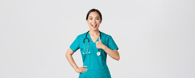 Covid-19, gezondheidswerkers, pandemisch concept. Glimlachende vrolijke, zelfverzekerde vrouwelijke aziatische verpleegster in scrubs hebben alles onder controle, duimen op als goedkeuring, garanderen de kwaliteit van de dienstverlening in de kliniek