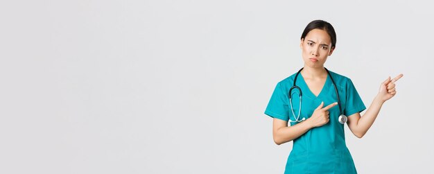Covid-19, gezondheidswerkers, pandemisch concept. Teleurgestelde en boze aziatische vrouwelijke therapeut, verpleegster in scrubs die boos naar de camera kijkt, mokkend en wijzend naar de rechterbovenhoek, wacht op uitleg.