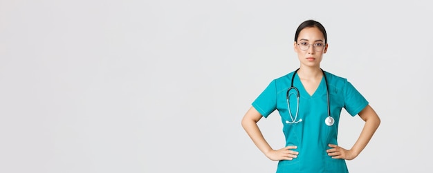 Covid-19, gezondheidswerkers, pandemisch concept. zelfverzekerde vastberaden aziatische vrouwelijke verpleegster helpt patiënten, werkt met coronavirusziekte, ziet er klaar uit, draagt scrubs en bril, witte achtergrond.