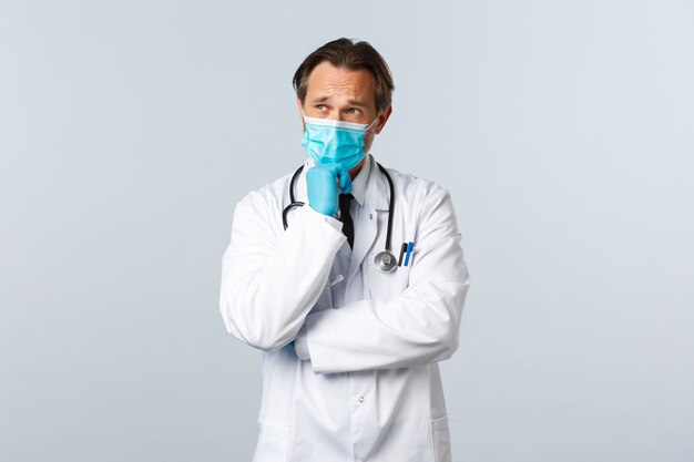 Covid-19, preventie van virussen, gezondheidswerkers en vaccinatieconcept. Doordachte serieuze mannelijke arts met medisch masker en handschoenen die denken, een keuze maken of een besluit nemen, witte achtergrond.
