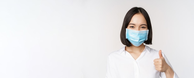 Covid en gezondheidszorgconcept Portret van een Aziatische vrouw die een medisch gezichtsmasker draagt en duimen laat zien die op een witte achtergrond staan