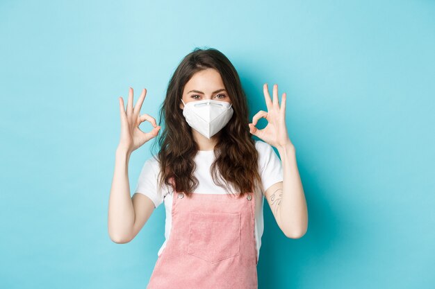 Covid, gezondheid en pandemisch concept. Tevreden mooi meisje in gasmasker, medisch masker met goed teken in goedkeuring, uisng maatregelen tegen coronavirus, blauwe achtergrond.