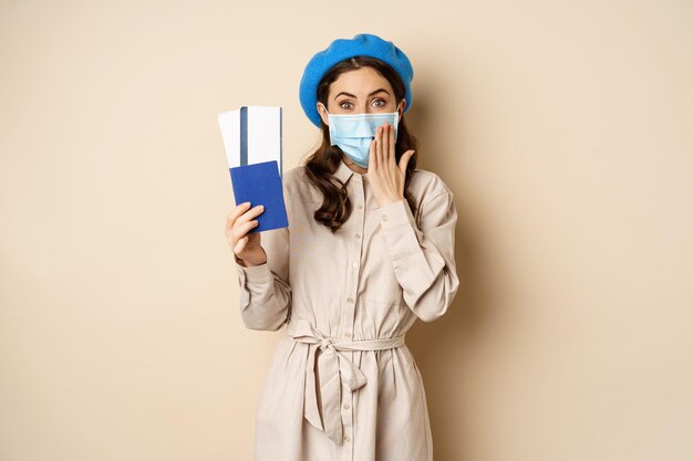 Covid pandemie en reizend concept. Portret van een schattig meisje met een medisch gezichtsmasker dat op reis gaat, paspoort met kaartjes in het buitenland laat zien en er opgewonden, beige achtergrond uitziet.
