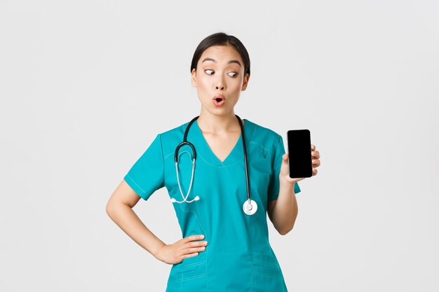 Covid19 gezondheidswerkers en online geneeskunde concept Opgewonden en verbaasde Aziatische vrouwelijke verpleegster arts kijken verrast terwijl ze een mobiele telefoon scherm internet consultatie app witte achtergrond toont