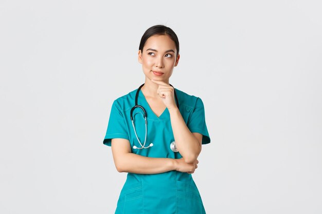 Covid19 gezondheidswerkers pandemie concept Doordachte slimme Aziatische verpleegster in scrubs wegkijken en denken glimlachend tevreden Dokter hebben interessant idee nadenken over witte achtergrond