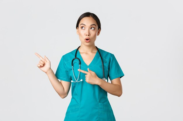 Covid19 gezondheidswerkers pandemisch concept Geïntrigeerde en verbaasde mooie aziatische vrouwelijke stagiaire verpleegster in scrubs of chirurg wijzend en kijkend naar de linkerbovenhoek en zeggend wow in verbazing