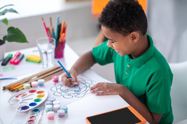 Creatieve activiteit. Profiel van een lachende jongen met een donkere huid in een groen t-shirt tekenen met een blauw potlood aan tafel in een lichte kamer