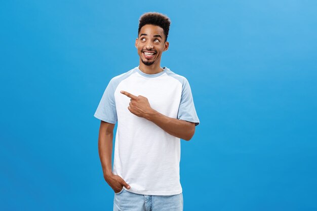 Creatieve afro-amerikaanse man met baard en afro-kapsel wijzend en starend naar de linkerbovenhoek, blij en tevreden met perfect weer en geweldig gezelschap over blauwe achtergrond