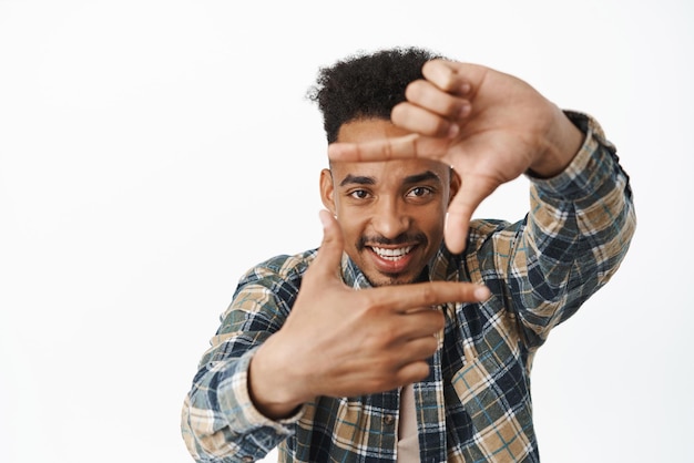 Creatieve afro-amerikaanse mannelijke student die door vingerframes kijkt en glimlacht, geamuseerd vastlegmoment neemt fotocamera die iets afbeeldt op een witte achtergrond