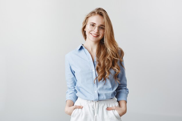 Creatieve kantoormedewerker ontspannen na een lange dag op het werk. Positieve knappe vrouw met blond haar in blauwe blouse, hand in hand in de zakken en glimlachend, vertrouwen uitend over grijze muur