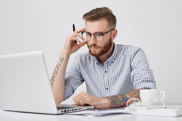 Creatieve mannelijke editor met tatoeages, kijkt zelfverzekerd naar het scherm van de laptop, werkt hard, omringd door moderne slimme telefoons