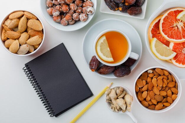 Dadels, amandelen, noten in witte borden met citroenachtige thee, gember, citrusvruchten, potlood en notitieblok plat op een witte tafel