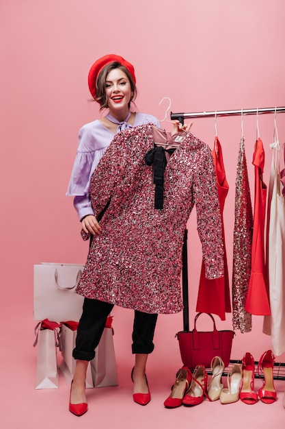 Dame in broek en blouse houdt jurk met pailletten. Vrouw poseren met pakketten tijdens het winkelen op roze achtergrond.