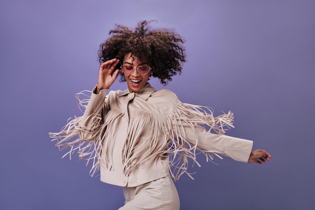 Dame in gefranjerde jas springen op paarse achtergrond Portret van brunette krullend vrouw in beige outfit dansen op geïsoleerde backdrop