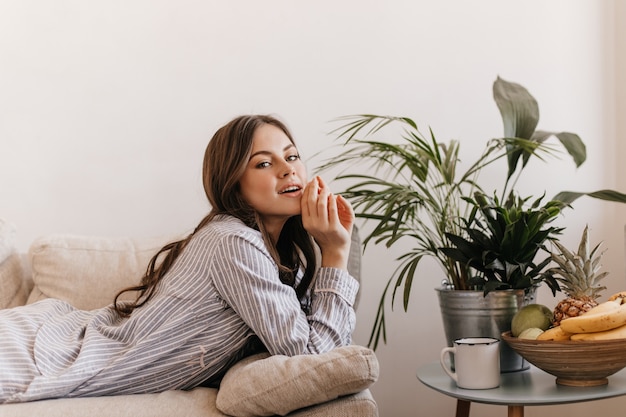 Gratis foto dame in gestreepte pyjama's die in woonkamer rusten. vrouw ligt op de bank naast fruitschaal