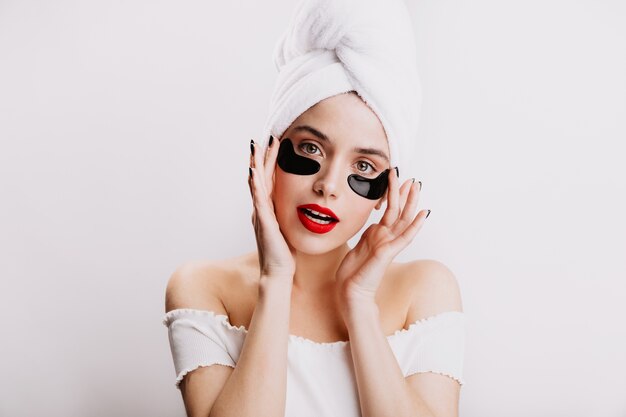 Dame in witte handdoek op haar hoofd poseert met zwarte vlekken voor oog. Meisje met rode lippenstift houdt van spa-behandelingen in de ochtend.