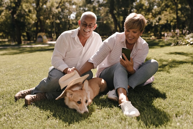 Dame met blonde haren in gestreepte blouse en spijkerbroek foto van hond maken en zittend op het gras met oude man in wit overhemd in park.
