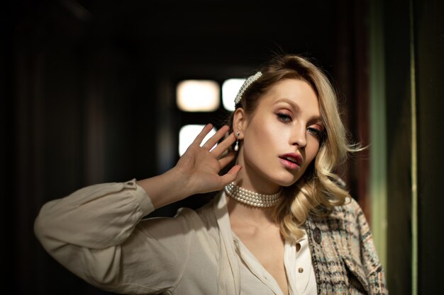 Dame met mooie make-up raakt oor aan. Mooie blonde vrouw in witte blouse en geruit jasje kijkt in de camera in de donkere kamer