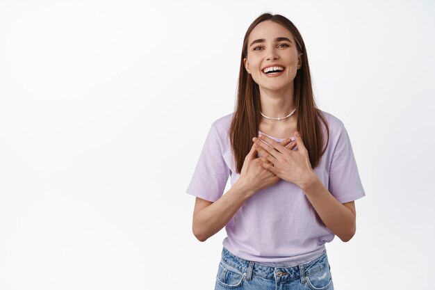 Dank je wel zo vleiend. Mooie jonge vrouw voelt zich dankbaar, glimlacht en houdt handen op de borst, drukt dankbaarheid en positieve emoties uit, staande op een witte achtergrond