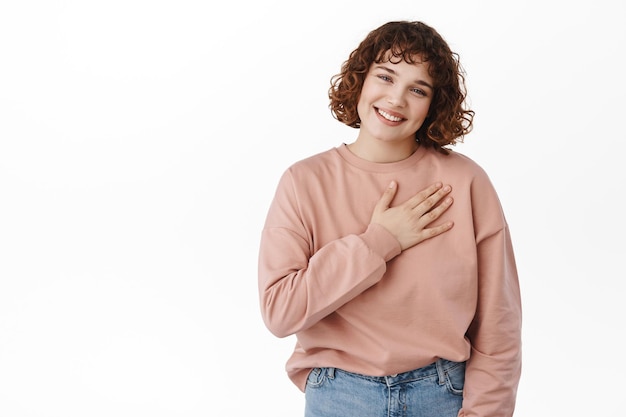 Dankbaar vriendelijk meisje, houd de hand op het hart en glimlach ontroerd, waardeer een aardig gebaar, bedankt voor iets, wees dankbaar, staande tegen een witte achtergrond.