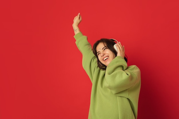 Dansen in een koptelefoon. Portret van de blanke vrouw geïsoleerd op rode muur met copyspace. Mooi vrouwelijk model in groene hoodie. Concept van menselijke emoties, gezichtsuitdrukking,