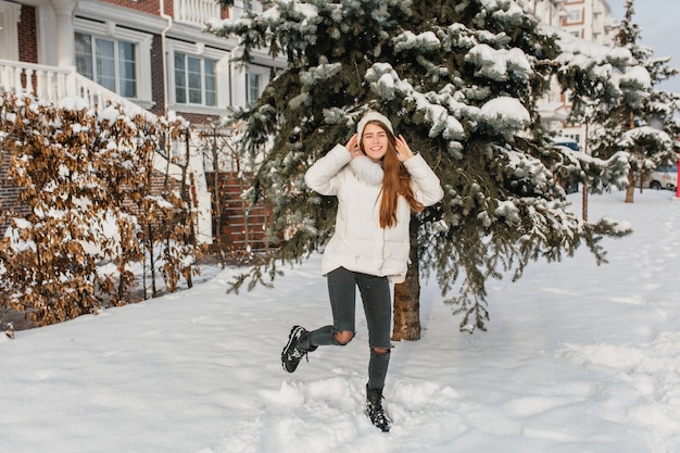 Dansende blanke vrouw in schattige gebreide muts. buiten portret van volledige lengte van zalige langharige dame in gescheurde broek die ronddoolt in de buurt van een besneeuwde boom.