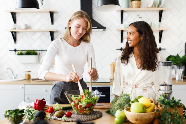 Gratis foto de aantrekkelijke kaukasische vrouw kookt gezonde salade en de mooie mulatvrouw kijkt op haar gekleed in zijdeachtige nachtjapon op modern ontworpen keuken