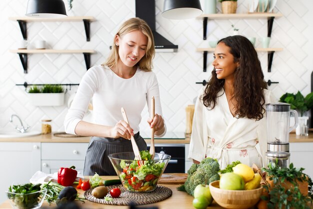 De aantrekkelijke Kaukasische vrouw kookt gezonde salade en de mooie mulatvrouw kijkt op haar gekleed in zijdeachtige nachtjapon op modern ontworpen keuken