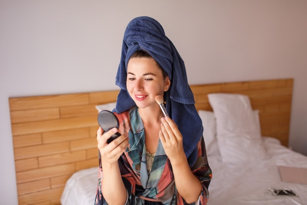 De aantrekkelijke vrouw die badjas, douchedoek op hoofd draagt treft voorbereidingen om op te maken, borstelt ter beschikking
