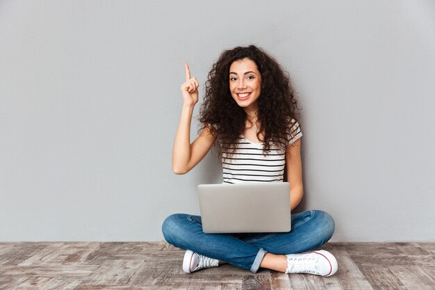 De aardige vrouw met mooie glimlach die opgewonden zijn om nuttige informatie in Internet via zilveren computer te vinden die eurekazitting in lotusbloem gesturing stelt op de vloer