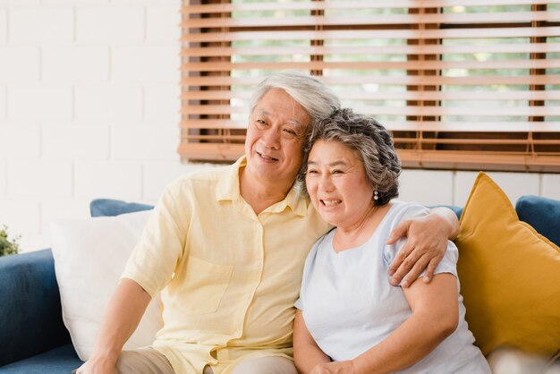 De Aziatische bejaarde paar het letten op televisie in woonkamer thuis, zoete paar geniet liefde van ogenblik terwijl het liggen op de bank wanneer ontspannen thuis.