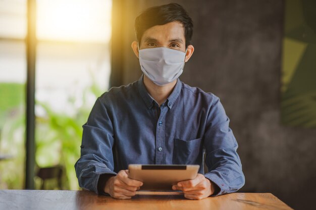 De aziatische mens die tablet gebruiken online werkend een dragend gezichtsmasker beschermt levensstijl nieuwe normaal van het coronavirus, mens levend in koffie binnen