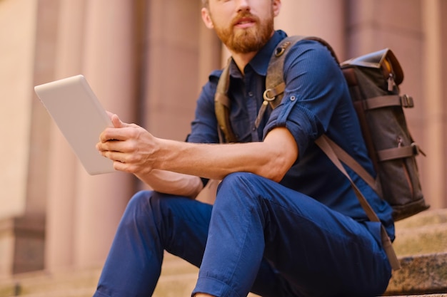 De bebaarde casual reiziger man zit op een trede en met behulp van een tablet-pc.