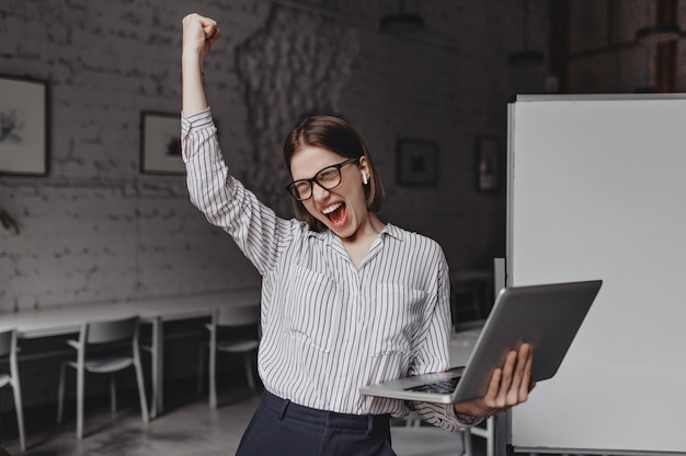 Gratis foto de bedrijfsvrouw met in hand laptop is tevreden met succes. portret van een vrouw in glazen en gestreepte blouse enthousiast schreeuwen en winnende gebaar maken.